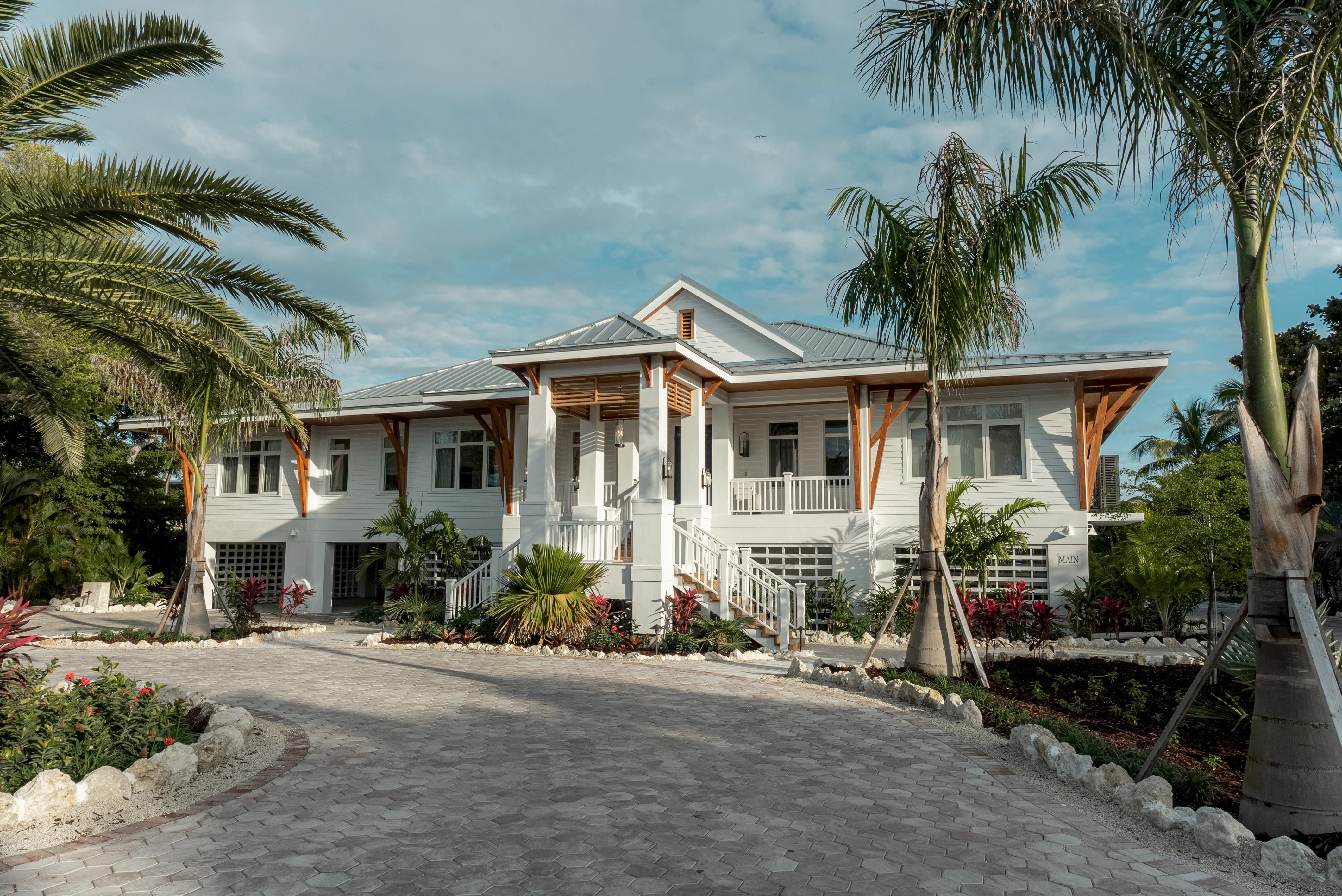 A large, light-colored bungalow with a wraparound porch, palm trees, a brick-paved driveway, and tropical landscaping under a sunny sky.