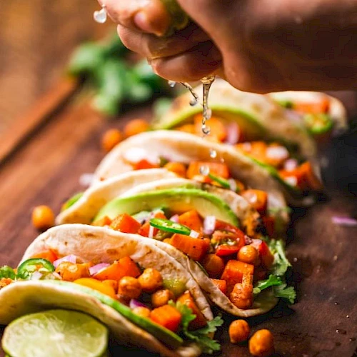 A person squeezes lime over a row of tacos filled with chickpeas, avocado, and vegetables on a wooden board, with lime slices nearby.