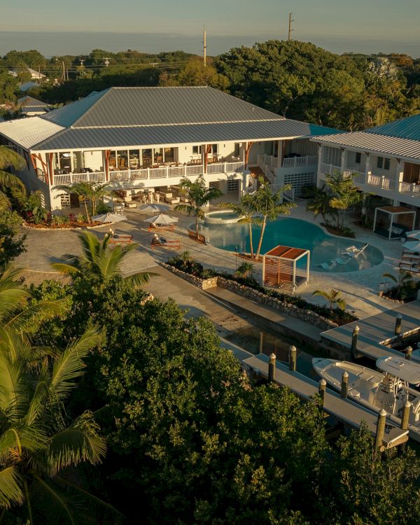 An aerial view of a tropical resort with two white buildings, a central pool, palm trees, and a wooden deck by the water, sunny and serene.
