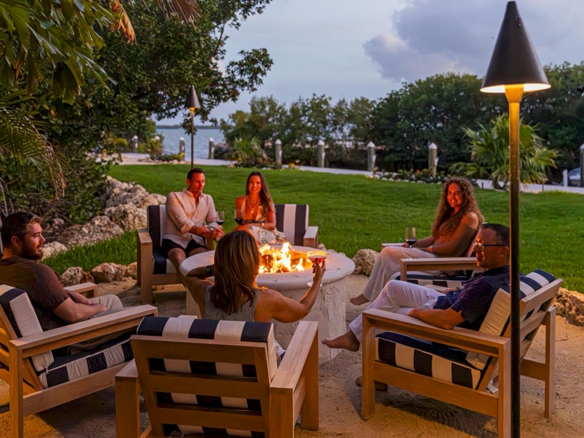 Friends lounge on a beach lawn around a crackling firepit with drinks under patio lights and a dusky sky.