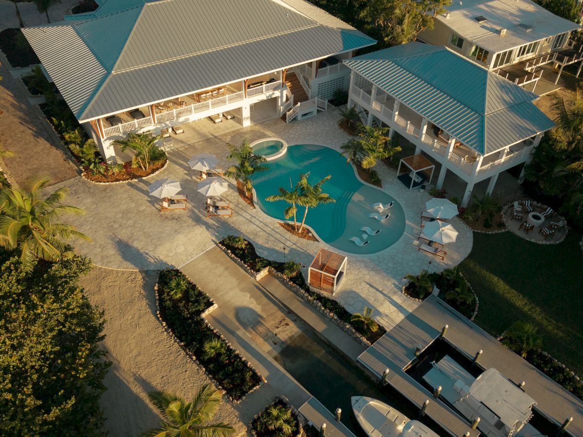 An aerial view of a tropical resort with turquoise pool, two white buildings with teal roofs, lounge chairs, palm trees, and a dock area.