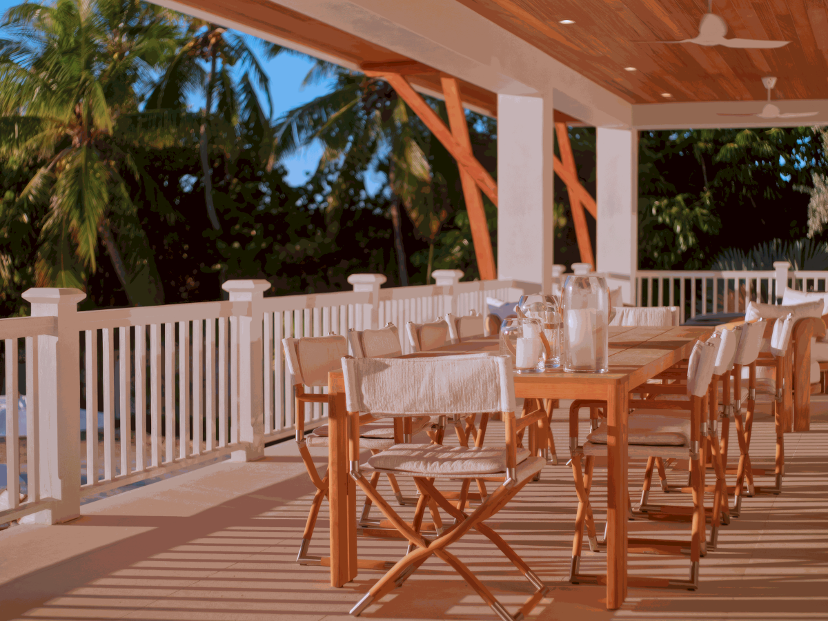 A sunlit veranda with a long dining table, wooden chairs, white tablecloths, glassware, and tropical trees in the background. End.