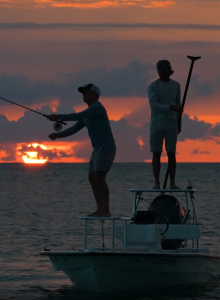 Two people on a boat at sunset, one lifting a hand as if posing, warm orange sky reflecting on calm water.