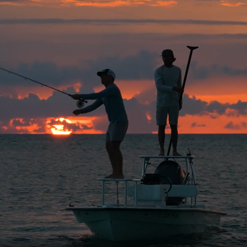 Two people on a small boat at sunset, one standing with arms raised, the other seated, silhouetted against a fiery orange sky.