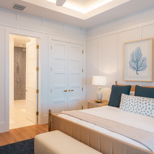 A bright hotel room with a large bed, blue and white accents, a desk with a mirror, a wall clock, and a window with sheer curtains.