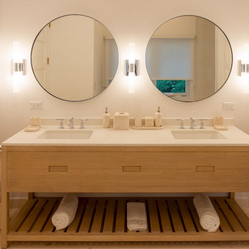 A modern bathroom with a wooden vanity, two sinks, large mirror, soft pendant lights, and a rounded wall mirror on the left.