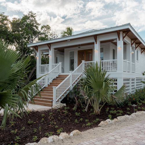 A small countryside house with a slanted roof, stone wall, potted plants, and a narrow pathway&mdash;cozy rustic home amid greenery.