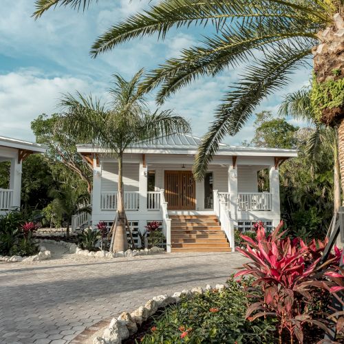 A sunny tropical house with a red-tiled roof, palm trees, and a white fence, inviting garden path and blooming flowers near a turquoise sky.