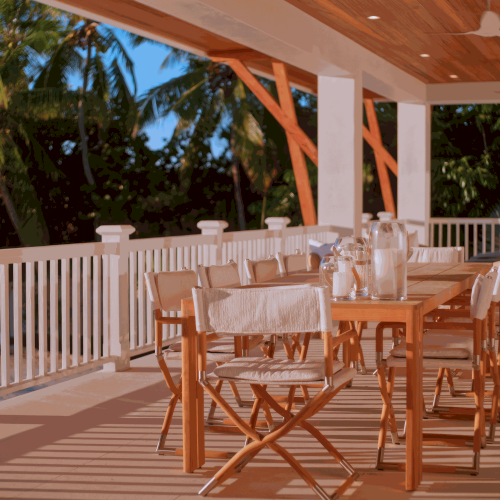 A wooden balcony with a white railing and chairs, sunlight, and a view of greenery in the background, under a wooden pergola.