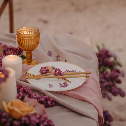 A beachside table setting with flowers, a candle, and a gold utensil set on a white plate, featuring a gold-rimmed glass.