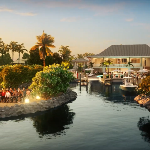 A waterfront wedding ceremony at sunset with a decorated arch, guests seated, surrounded by palm trees and a house in the background.