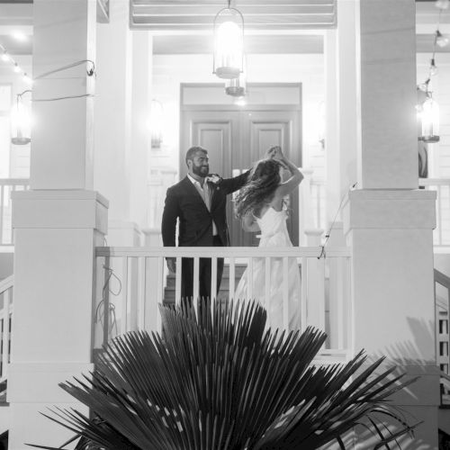 A man and woman stand on a porch balcony, the man touchingly adjusting her hair, framed by pillars and bright courtyard lights.