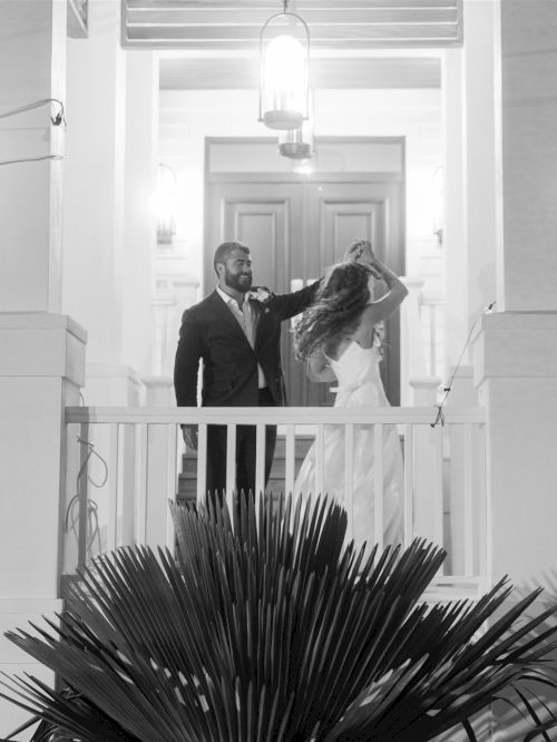 A man and woman stand on a porch balcony, the man touchingly adjusting her hair, framed by pillars and bright courtyard lights.