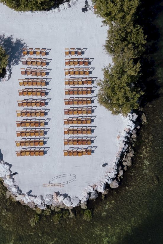 An aerial view shows a stone platform on water with two neat rows of lounge chairs facing the center, surrounded by green foliage and clear water.