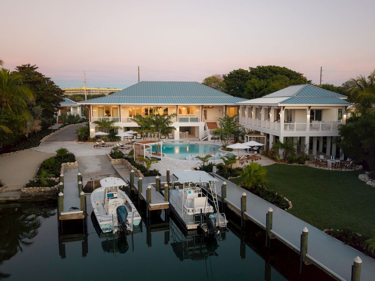 A waterfront resort with a central pool, two blue-roofed buildings, palm trees, and a dock with boats along calm canal at dusk.