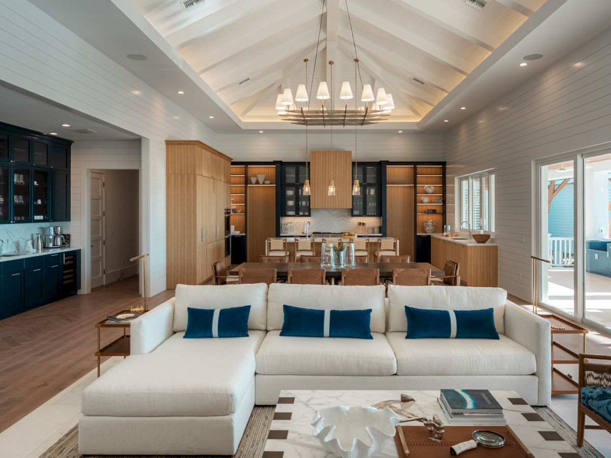 A bright, airy open-plan living and dining room with a white sofa, blue cushions, wooden accents, and a modern kitchen in the background, plus a chandelier.