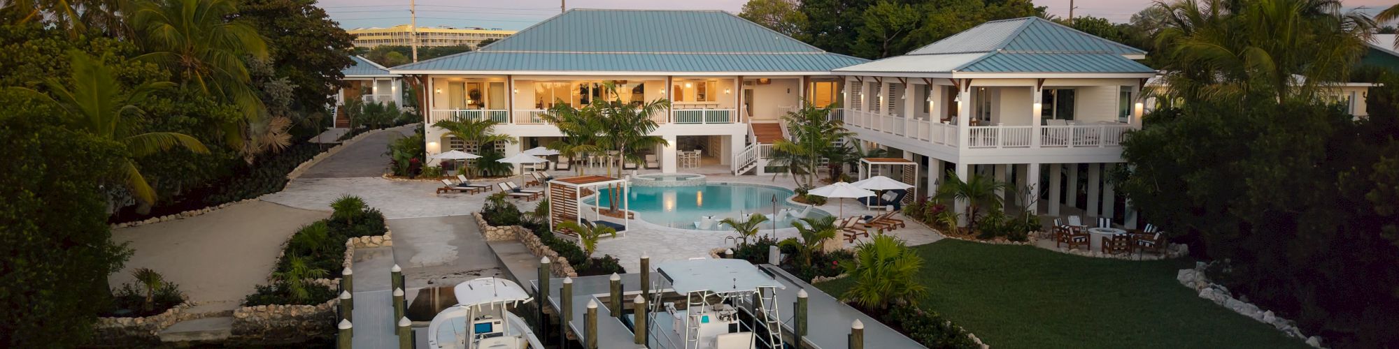 A luxurious waterfront villa with a pool, palm trees, a dock, and boats, overlooking calm water at sunset, surrounded by lush greenery.