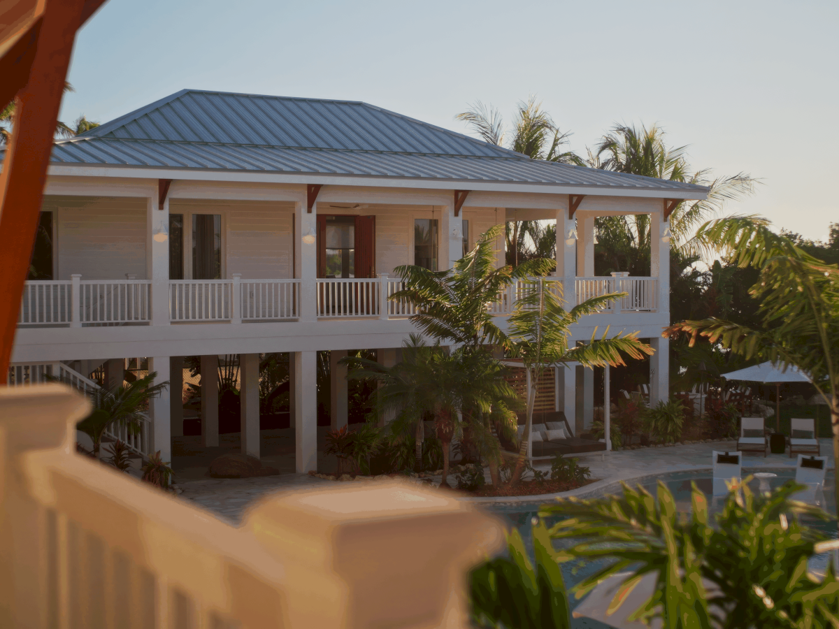 A two-story tropical resort with a blue metal roof, white railings, lush palm trees, and a sunlit swimming pool area.