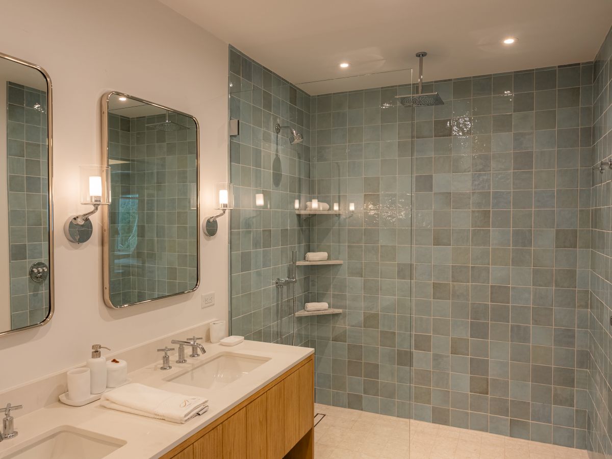 A modern bathroom featuring double sinks, a wooden vanity, two framed mirrors, wall sconces, and a tiled walk-in shower with multiple shelves.
