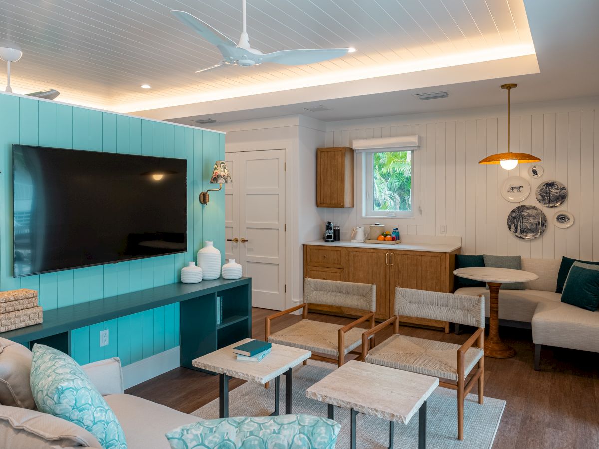 A bright, coastal living room with a turquoise entertainment wall, wall-mounted TV, wicker and wood furniture, and a cozy seating area near a small kitchen nook, all under soft natural light.
