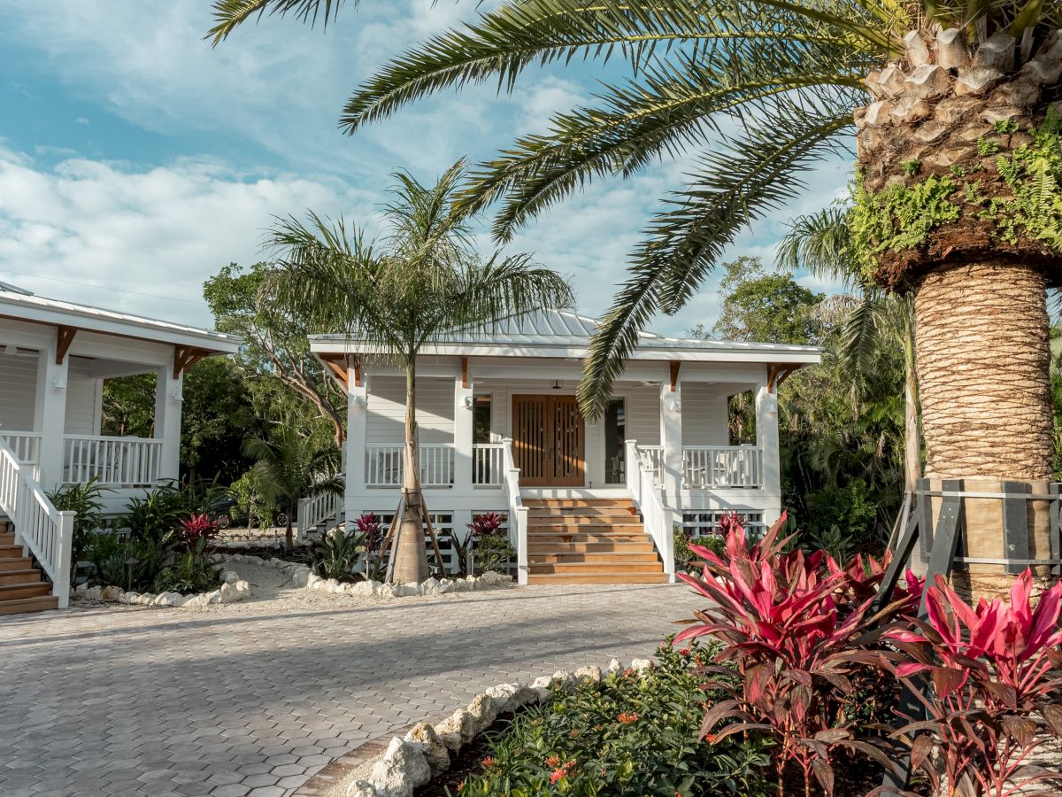 A row of cozy single-story cottages with shaded porches, tropical palm trees, and vibrant pink plants lining a paved walkway, sunny and inviting.