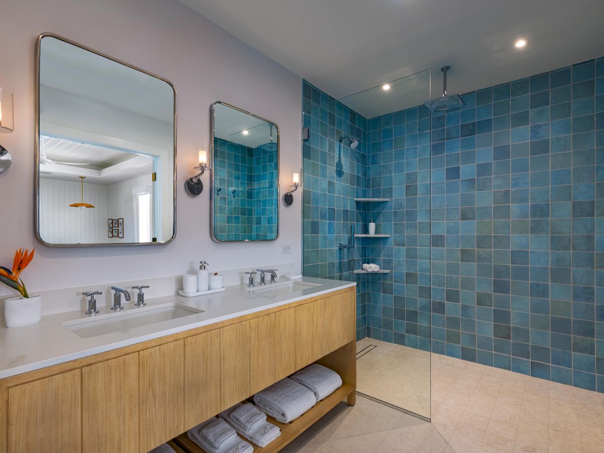 A modern shared vanity with three sinks, a long wooden cabinet, two mirrors, and a glass-enclosed blue-tiled shower area; towel shelves below.
