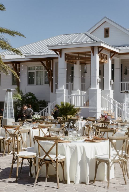Outdoor patio with round tables, white tablecloths, wooden chairs, and a white house-style building in the background under palm trees.
