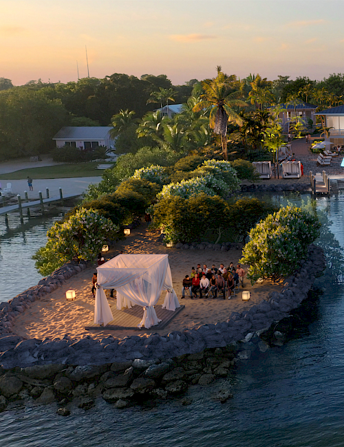 A tropical waterfront scene: palm trees, lush greenery, a small island with a tented seating area by the water, and a cozy resort at dusk.