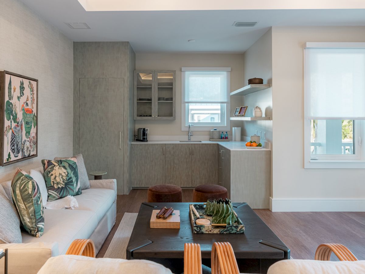 Cozy, modern living room with a light gray kitchen, white walls, and a large window; a sofa with patterned pillows faces a dark coffee table, stools by the counter, and wall art.