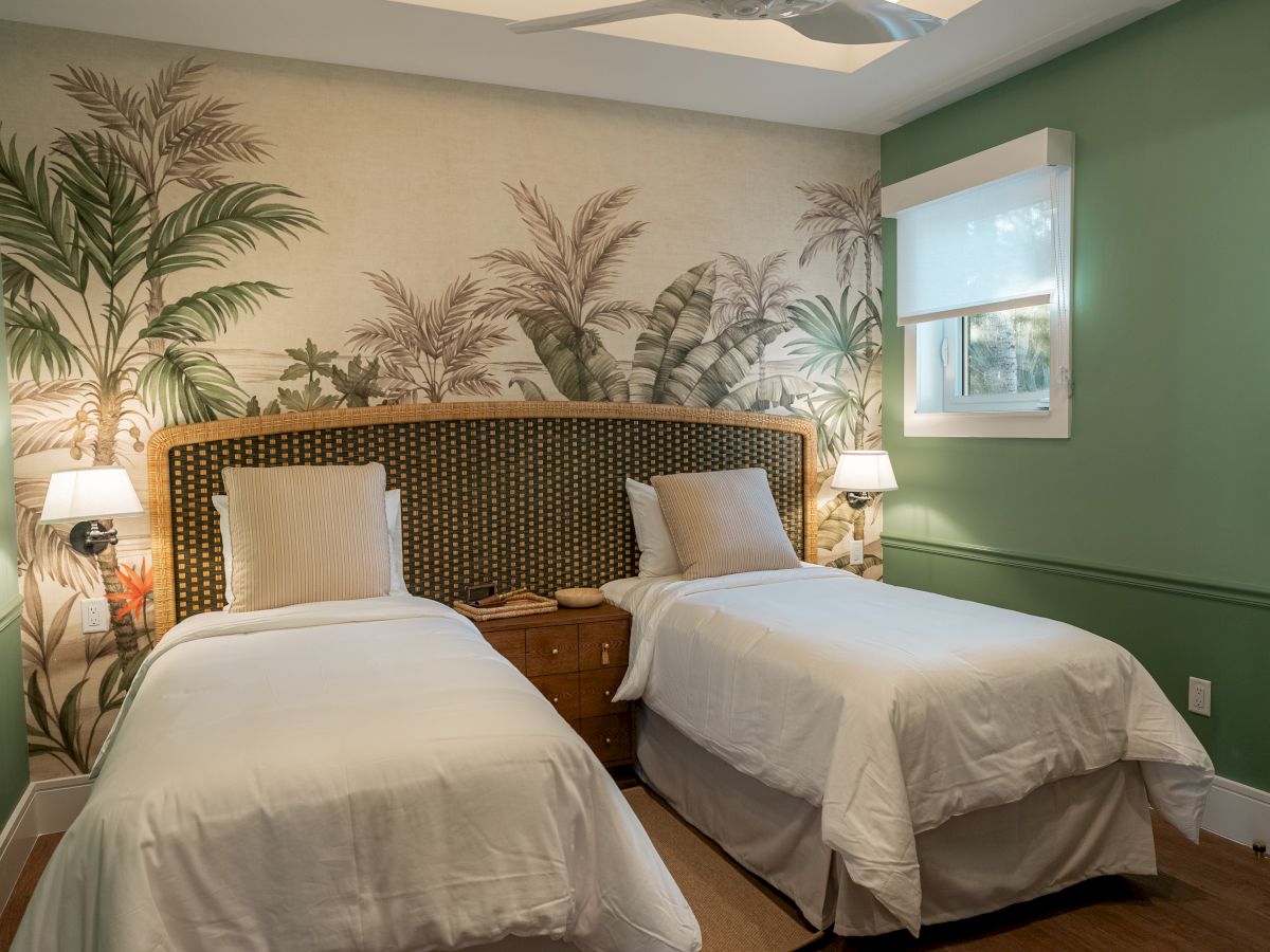 A twin bedroom with two single beds, a shared wooden headboard, botanical wall mural, green walls, nightstands, and lamps on each side.