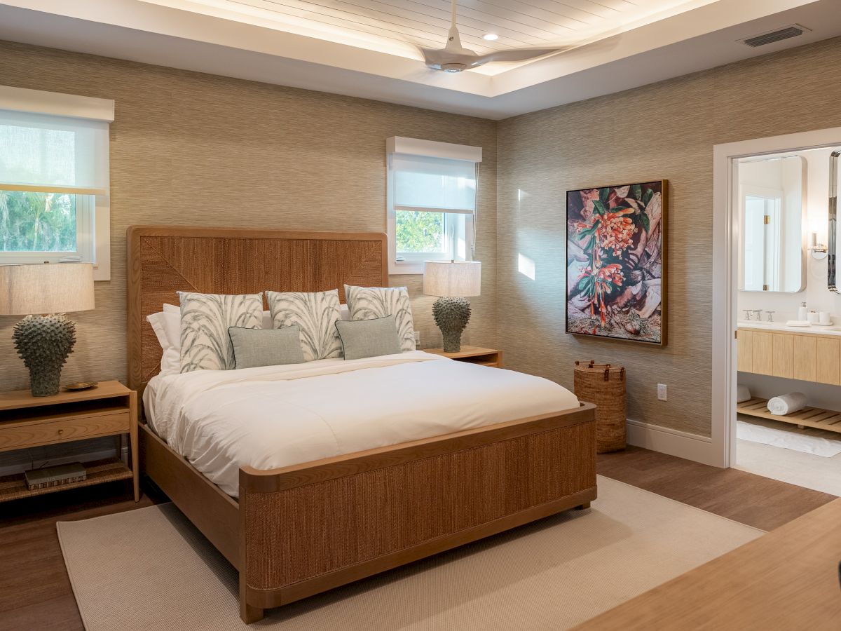 A cozy, modern hotel bedroom with a wooden king bed, two nightstands, soft lighting, and a doorway to a bright ensuite; neutral tones and a framed artwork.