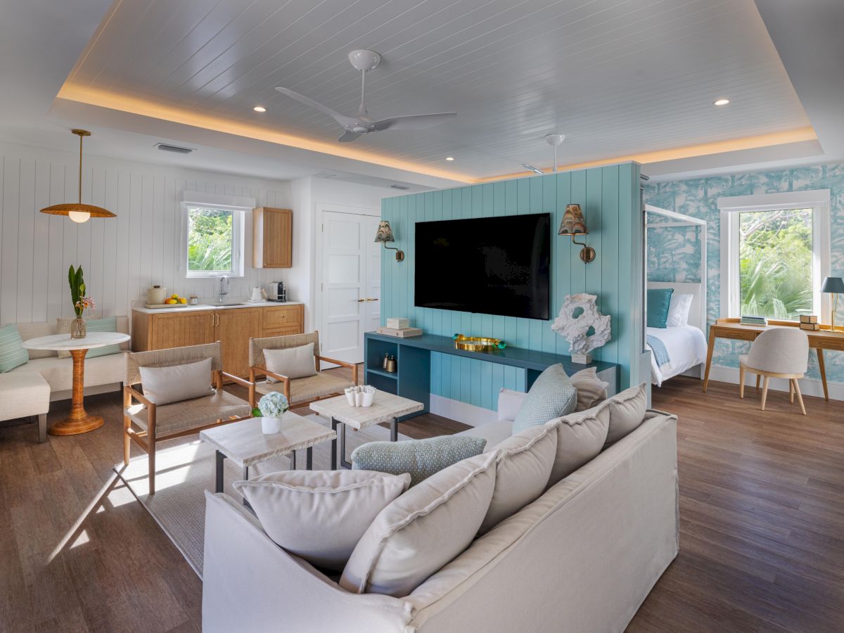 A bright, open living space with a white and beige color scheme, blue feature wall with a TV, comfy sectional, mid-century chairs, a wooden kitchen area, and large windows letting in natural light.