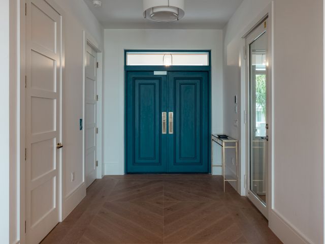 A bright foyer with white walls, a bold teal double front door, tan herringbone floor, and side panels/windows flanking the entry.Top it at 140 characters, always ending the sentence.