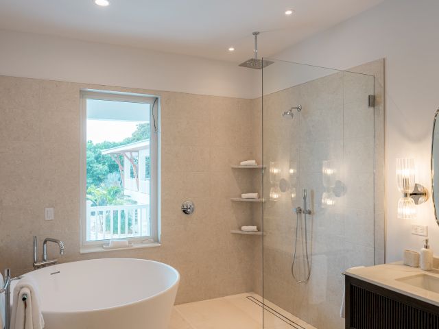 A modern bathroom with a freestanding tub, glass-walled shower, beige tiles, a window, and a vanity with a round mirror, all in a bright, spa-like setup.