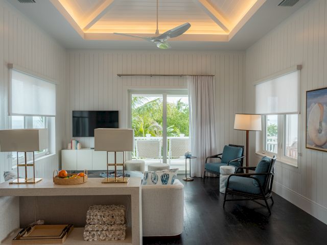 A bright, coastal living room with white paneled walls, a white sofa, dark wood floors, a ceiling fan, and large windows/doors opening to greenery, plus chairs, lamps, and a wall-mounted TV. Top it at 140 characters, always ending the sentence.