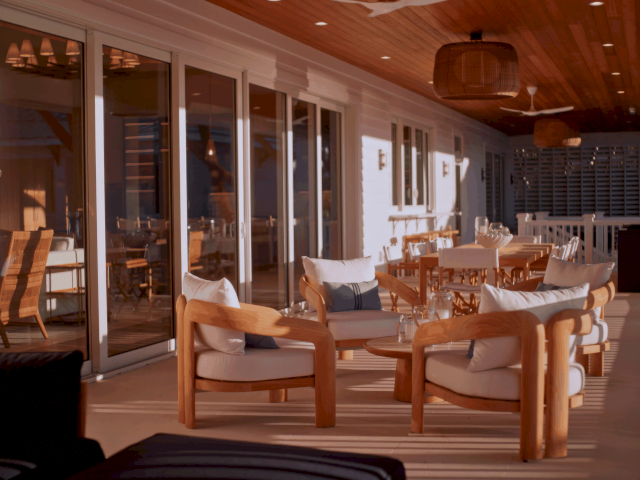 A cozy, sunlit living room with a wooden ceiling, warm seating, a coffee table, and a sideboard; inviting homey space with shelves and plants.