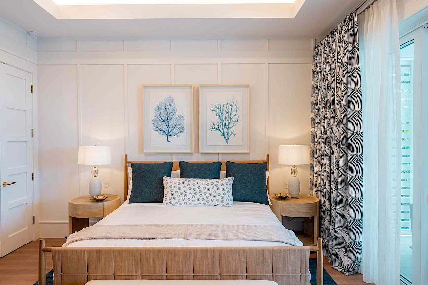 A bright, modern bedroom with a white and beige color scheme, matching lamps, two framed botanical prints, and blue accent pillows on a neatly made bed.