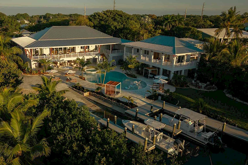 Aerial view of a waterfront villa with a pool, multiple decks, palm trees, and boats along a canal, set in a tropical coastal neighborhood.