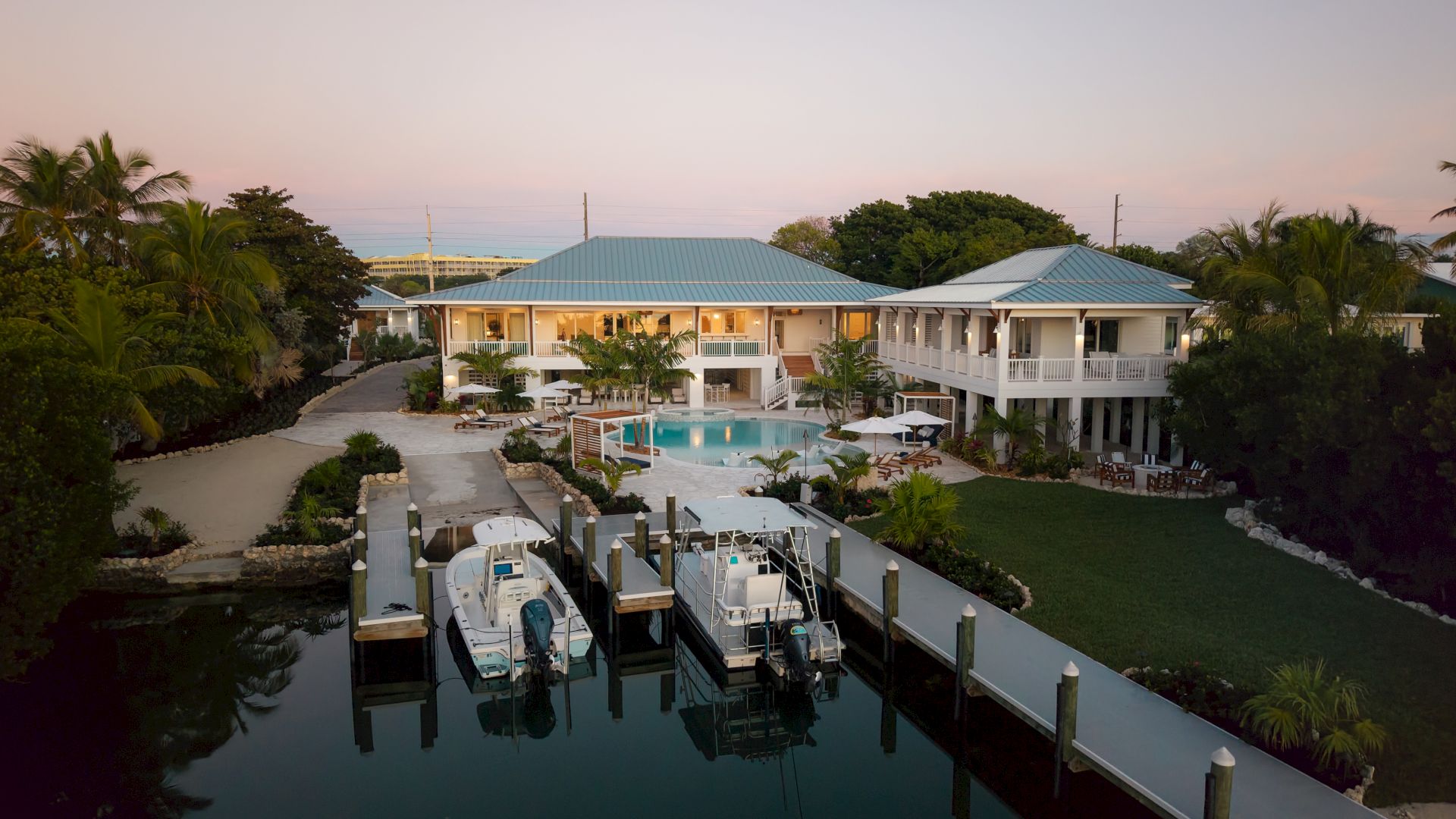 A tropical resort with a two-story white building, blue roofs, a central pool, and a dock with boats along calm water.