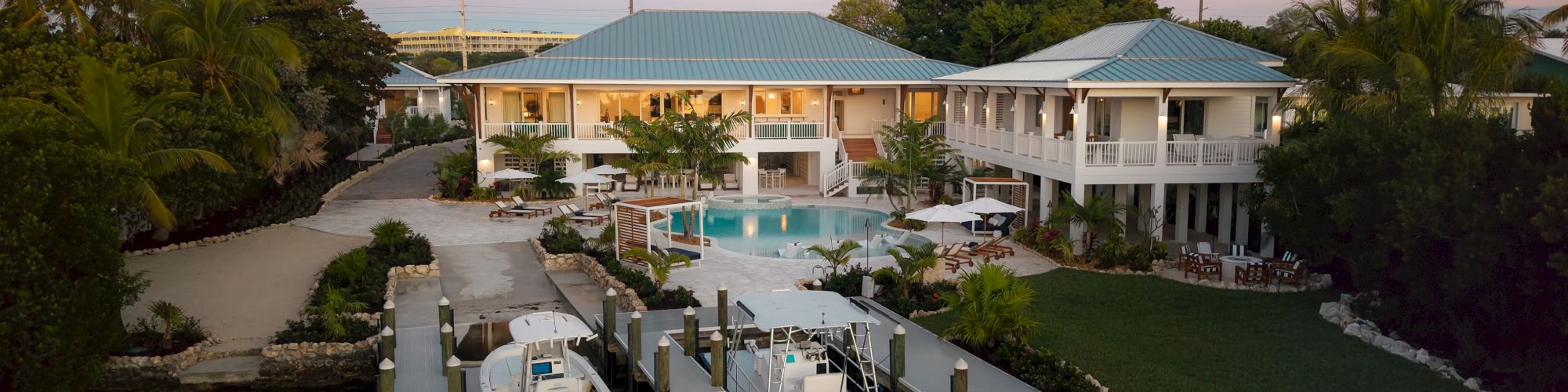 A tropical resort with a two-story white building, blue roofs, a central pool, and a dock with boats along calm water.