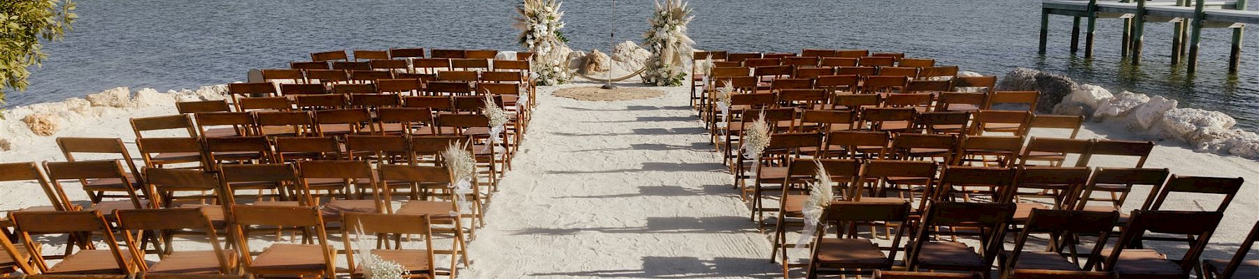 Beach wedding setup with wooden chairs, a floral arch on white sand, and calm ocean in the background.