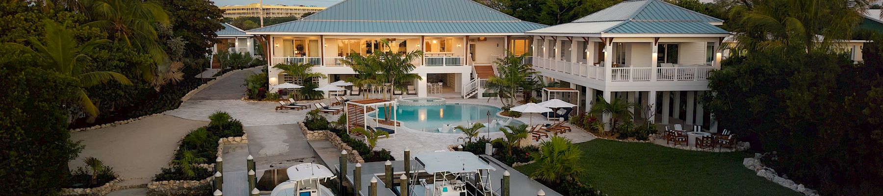A tropical seaside mansion with two blue-roofed pavilions, a central pool, docks, and a boat-filled marina at sunset.
