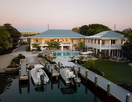 A tropical seaside mansion with two blue-roofed pavilions, a central pool, docks, and a boat-filled marina at sunset.