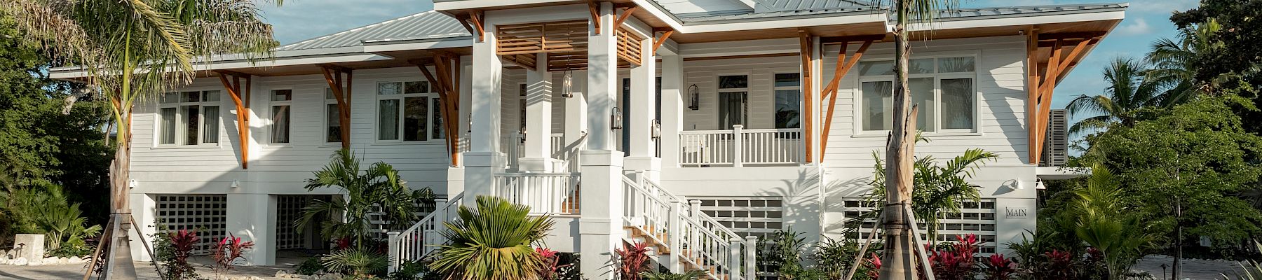 A single-story white house with a wraparound porch, palm trees, and a curved paved driveway under a blue sky, tropical garden surrounding it.