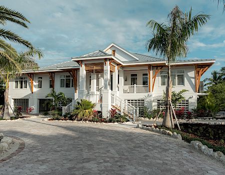 A single-story white house with a wraparound porch, palm trees, and a curved paved driveway under a blue sky, tropical garden surrounding it.